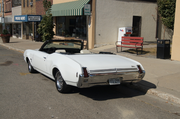 Ein weißes Auto mit der Nummer 771-299 steht auf der Straße, daneben eine Bank, im Hintergrund ein Gebäude und ein Geschäft namens "Immobilienversicherung".