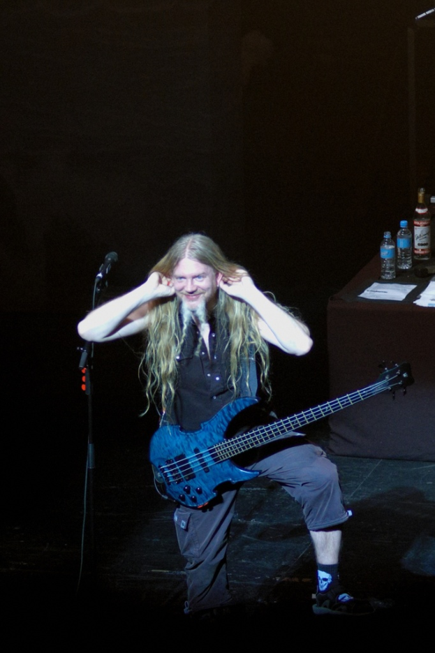 Ein Mann steht und lächelt, trägt eine blaue Gitarre und hält ein Mikrofon auf einem Ständer; auf einem schwarzen Tuch liegt ein Tisch mit Wasserflaschen, Papieren und einer Weinflasche vor einem dunklen Hintergrund.
