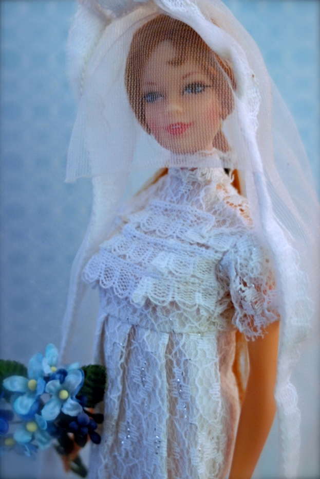 Eine Puppe in einem Kleid, Schleier und mit einem Strauß, vor einem Hintergrundobjekt.