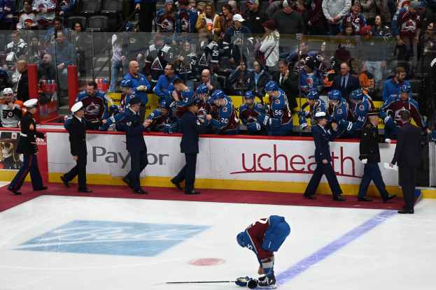 Ein Hockey-Spieler in dunkler Uniform hält einen Stock auf dem Eis, umgeben von Menschen in Mützen und mit Hockey-Stöcken in der Hand, sowie Zuschauern auf Sitzen und Stehplätzen im Hintergrund.