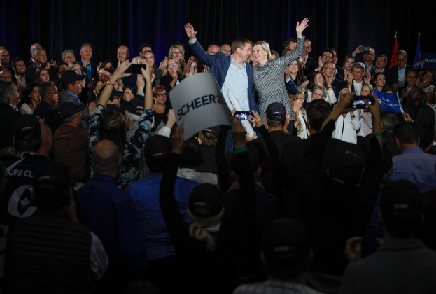 Ein Mann und eine Frau stehen vor einer Menge, einige halten Schilder und Mobiltelefone, sowie Fahnen und einen Vorhang im Hintergrund.