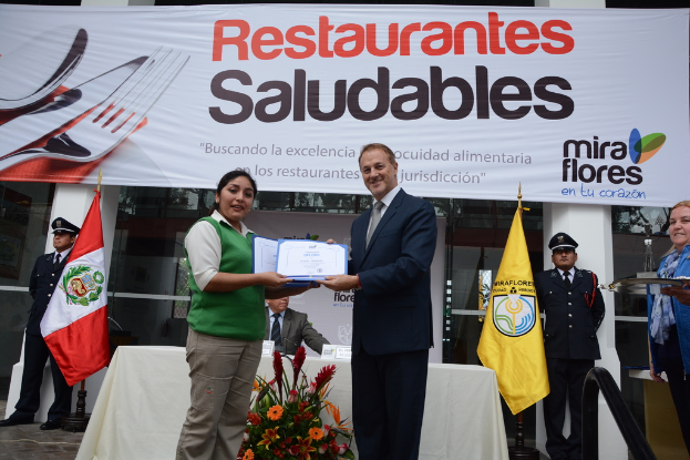 Eine Frau und ein Mann lächeln, während sie Zertifikate halten und vor einem Tisch mit dekorativen Blumen, einem Stab und einem Tuch stehen, auf dem verschiedene Gegenstände liegen und im Hintergrund Flaggen, Menschen, Banner, Säulen, Fenster und Glasobjekte zu sehen sind; eine Person rechts hält ein Tablett und lächelt.