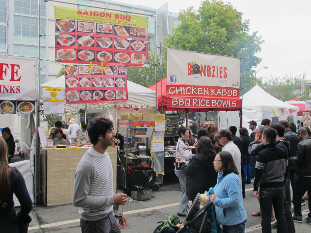 Menschen stehen auf einer Straße außerhalb der Stadt, mit einem Mann in einem grauen und weißen T-Shirt, der eine Wasserflasche und eine Frau in einer blauen Jacke, die einen Kinderwagen schiebt. Im Hintergrund sind Stände mit weißen und roten Zelten, farbige Banner und Gebäude, Bäume und eine Straßenlaterne zu sehen.