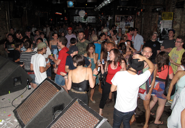 Eine Gruppe von Menschen, die tanzen und dabei einige Mützen tragen und Taschen haben, mit einer Soundbox, Kabeln, einer Flasche und Pfosten mit Text im Hintergrund.