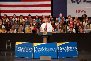 Ein Mann in einem weißen Hemd und roter Krawatte steht an einem Podium mit drei Texttafeln, einem Hocker mit Mikrofon und einer sitzenden Menge im Hintergrund, mit einer Flagge und zusätzlichem Text.