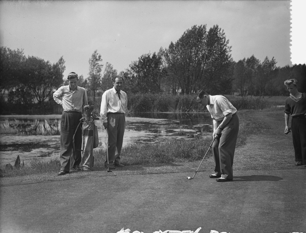 Eine Gruppe von Männern, die Golf auf einem grünen Kurs mit Bäumen und einem Wasserlauf in der Ferne spielen, in Schwarz-Weiß aufgenommen.
