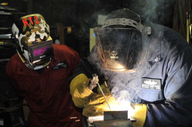 Zwei Schweißer in Helmen arbeiten an einem Metallstück in einer Werkstatt, mit Rauch vom Schweißen und verstreuten Werkzeugen.