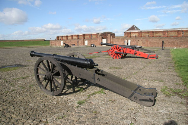 Kanonen auf Kopfsteinpflaster im Vordergrund mit einer Festungsmauer links, unter einem teilweise bewölkten, sonnigen Himmel.