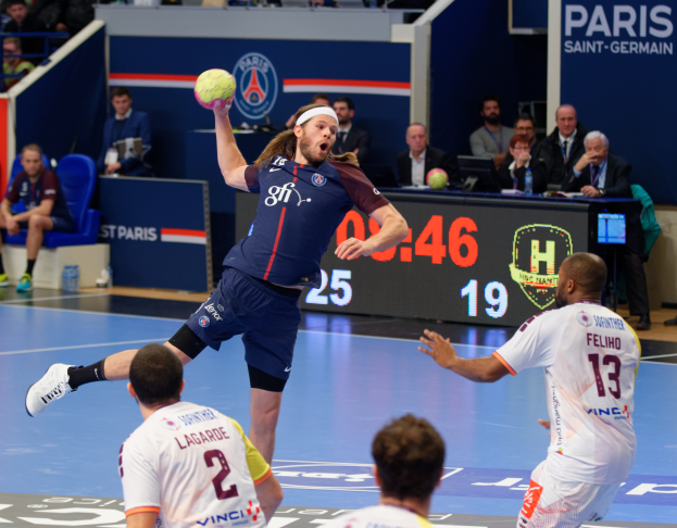 Männer spielen Handball auf einem Feld mit einem Ball in der Luft, Zuschauer sitzen und stehen im Hintergrund, ein Tisch mit einem Monitor und Flaschen sowie eine Tafel mit Text und Logos, die ein Spiel zwischen Paris Saint-Germain und Olympique Lyon anzeigen.