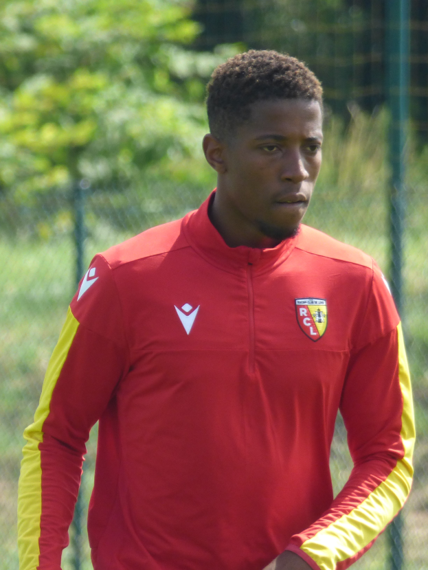 Ein junger Mann in einem roten und gelben Trikot steht auf einem Fußballfeld, umgeben von einem Zaun und Bäumen, mit einem entschlossenen Gesichtsausdruck, der wie ein Flügelstürmer aussieht.