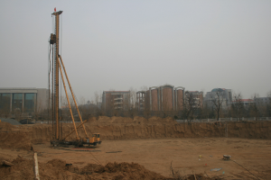 Eine Baustelle mit einem Kran in der Mitte, umgeben von Gebäuden, Bäumen und einem klaren blauen Himmel, mit Erde bedecktem Boden und verstreuten Gegenständen.
