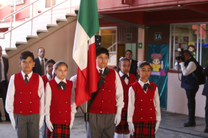 Eine Gruppe von Kindern in Schuluniformen steht zusammen, eines hält eine Fahne, mit Menschen, einer Kamera, einem Gebäude, einem Schild, einer Fahne und einer Treppe im Hintergrund.