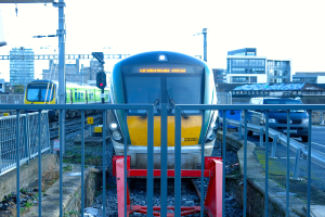 Zwei Züge auf parallelen Gleisen mit einem Zaun im Vordergrund, ein Auto auf der Straße rechts, eine Signalleuchte auf einem Pfahl und Gebäude im Hintergrund unter einem Himmel.