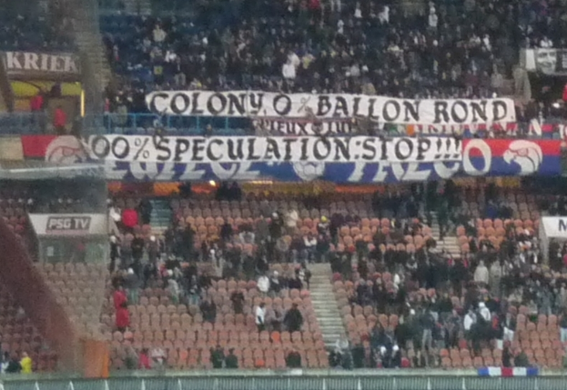 Ein Stadion mit einer großen Menschenmenge, Fahnen und Stühlen, einem Zaun unten und einer Treppe links.