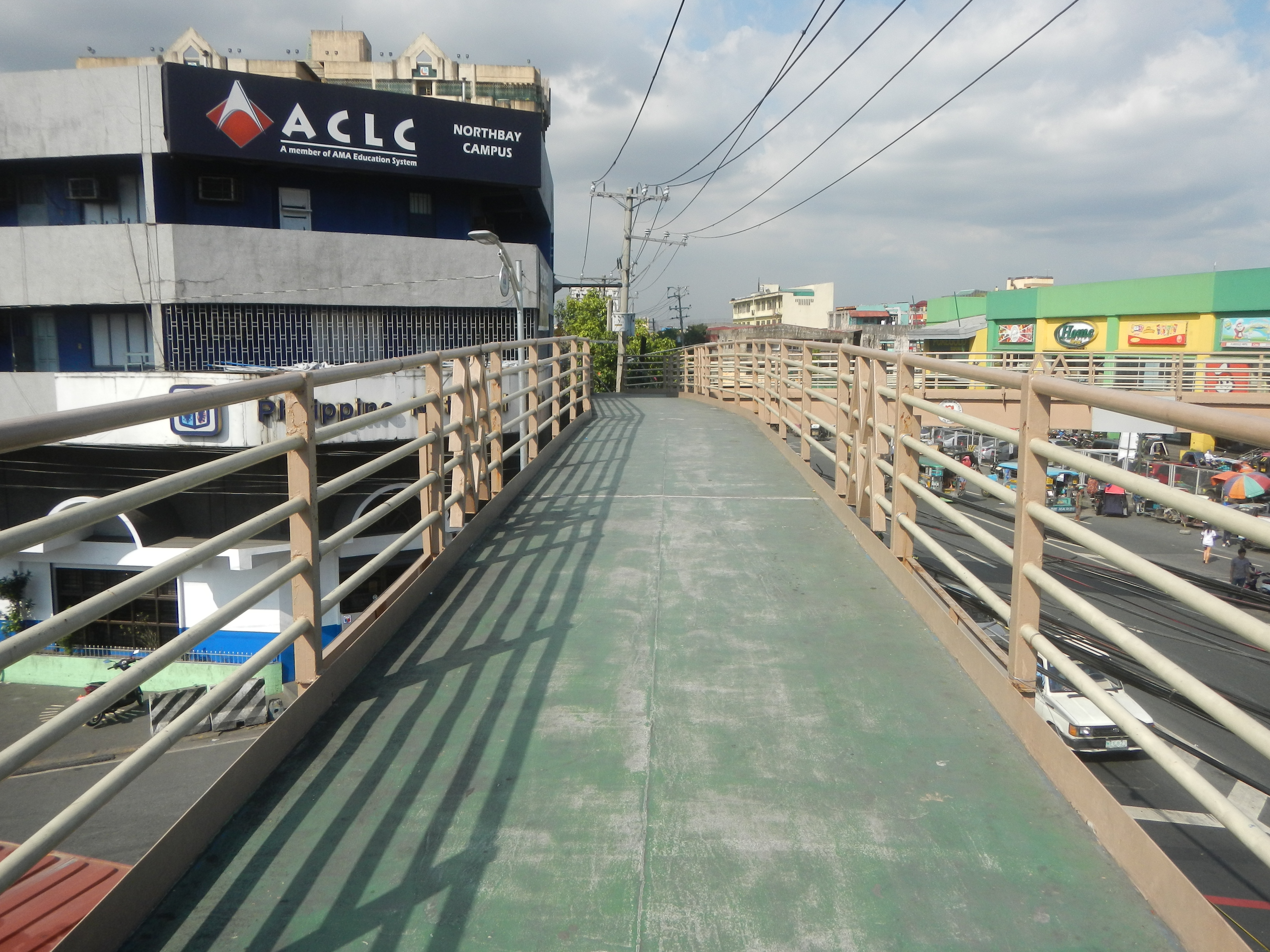 Eine Fußgängerbrücke mit Geländer überspannt eine Stadtstraße, darunter fahren Fahrzeuge; Gebäude mit Schildern flankieren die Brücke, und im Hintergrund sind Strommasten mit Drähten, Bäume und ein bewölkter Himmel zu sehen.