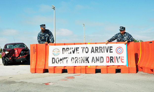 Zwei Polizeibeamte stehen neben einem beschädigten Auto mit einer Tafel davor, unter einem klaren blauen Himmel mit Laternen und Bäumen im Hintergrund.