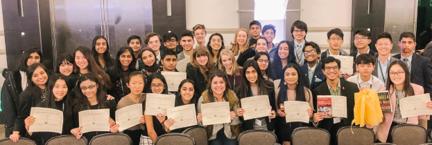 Gruppe von Menschen mit Urkunden, die auf einem Foto posieren, in einem Raum mit Stühlen und einer Wand im Hintergrund.