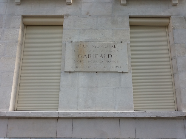 Ein Gebäude mit zwei Fenstern und Läden, das eine Gedenktafel an seiner Seite trägt.