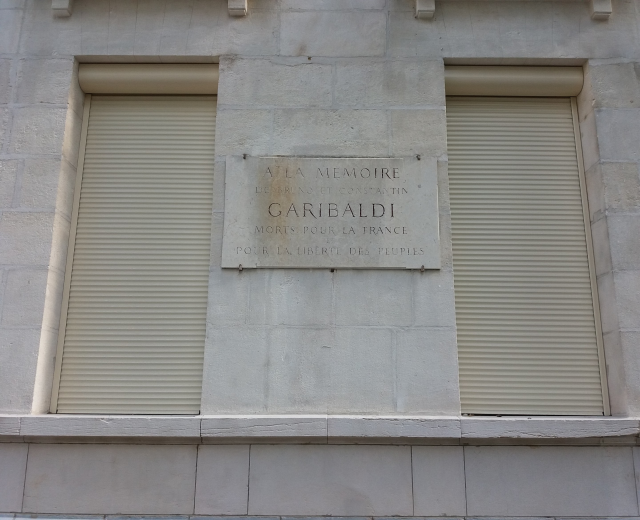 Ein Gebäude mit zwei Fenstern und Läden, das eine Gedenktafel an seiner Seite trägt.