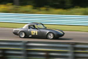 Ein Auto mit einem Helm tragenden Fahrer bewegt sich auf einer Straße, die von Barrieren gesäumt ist und Bäume im Hintergrund hat.