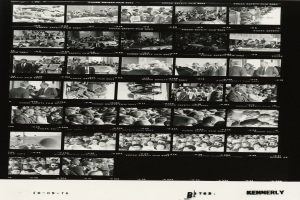 Schwarzes und weißes Foto einer vielseitigen Menge, die lachend auf einen Fernseher schaut.