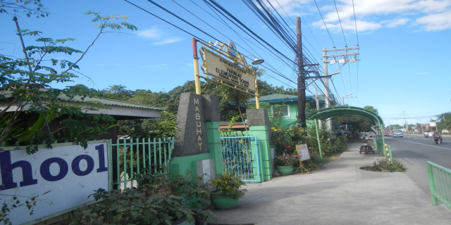 Einfahrt zu einer Schule mit einem "Schule"-Schild am Straßenrand, mit Fahrzeugen, einem Tor, einem Tor, einer Namensschild, Grünflächen, Strommasten mit Drähten und einem bewölkten Himmel im Hintergrund.