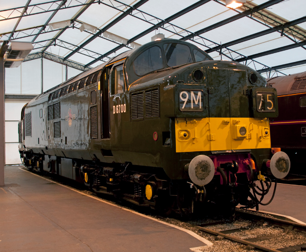 Ein Zug fährt auf den Schienen neben einem Bahnsteig mit einem am Dach befestigten Pfahl, der Metallstangen mit Lampen trägt.