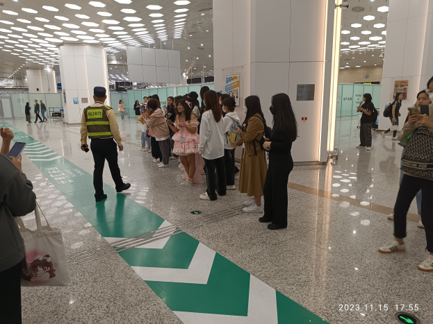 Eine Gruppe von Menschen steht in einer Reihe an einem Flughafen-Sicherheitscheck-in, einige halten Taschen und Mobiltelefone, mit Säulen im Hintergrund und Lampen an der Decke und einem Wasserzeichen am unteren Bildrand.