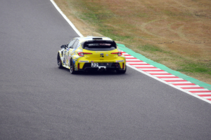 Ein gelber Sportwagen mit Text und Zahlen darauf fährt auf einer Rennstrecke, flankiert von Gras auf beiden Seiten.