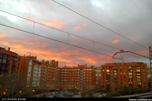 Gebäude mit Fahrzeugen und Bäumen davor, Strommast mit Kabeln oben und Himmel im Hintergrund.
