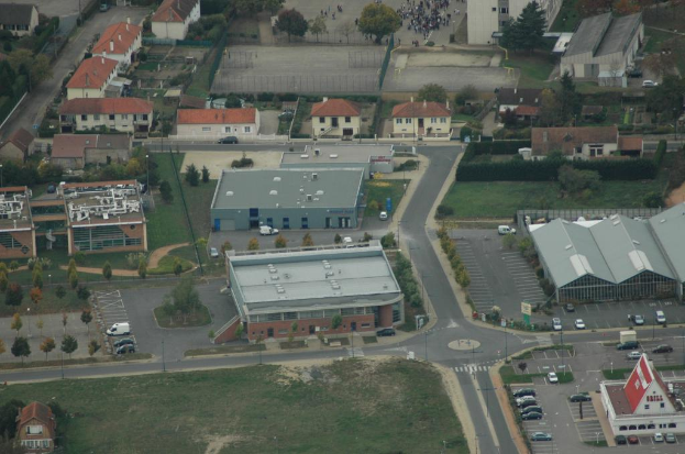 Luftaufnahme von dicht bebauten Gebäuden mit Straßen, parkenden Autos und Grünflächen dazwischen.