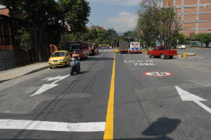 Eine Straßenszene mit Gebäuden, Bäumen, fahrenden Fahrzeugen, Schildern, Verkehrsampeln, einem blauen bewölkten Himmel und Warnschildern auf der Straße.
