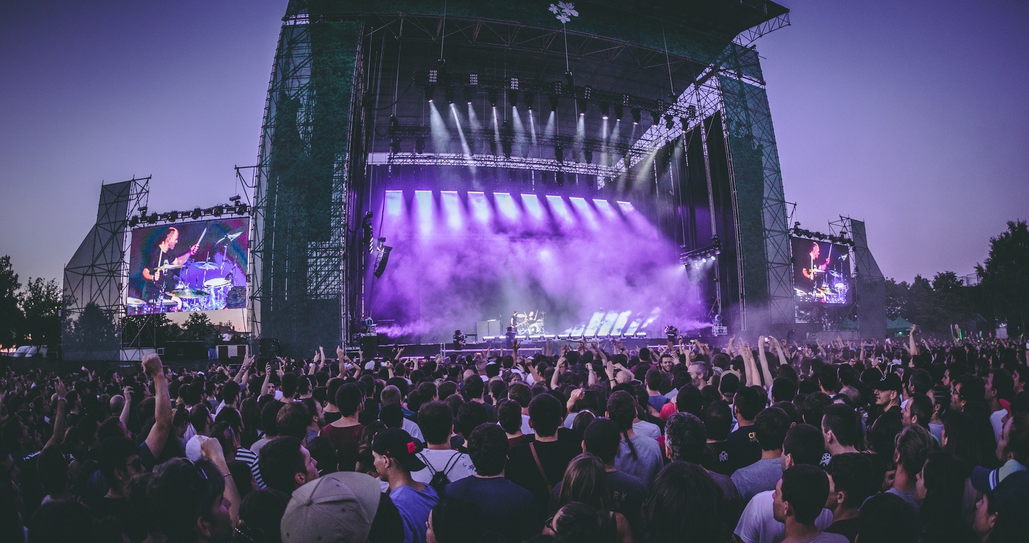 Eine große Menschenmenge schaut bei einem Musikfestival auf einer beleuchteten Bühne mit Instrumenten und Bildschirmen, die vor einem Gebäude, Bäumen und einem klaren blauen Himmel aufgebaut sind.