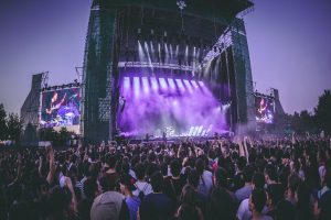Eine große Menschenmenge schaut bei einem Musikfestival auf einer beleuchteten Bühne mit Instrumenten und Bildschirmen, die vor einem Gebäude, Bäumen und einem klaren blauen Himmel aufgebaut sind.