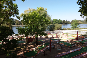 Ein Minigolf-Platz in einem Park mit grünem Gras, Bäumen und einem Zaun, mit Stühlen und Tischen, einem Gewässer und einem Boot im Hintergrund und einem sichtbaren Himmel.