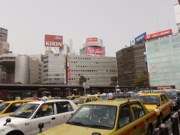 Eine Stadtansicht mit Fahrzeugen auf der Stra√üe, Geb√§uden, B√§umen, Masten mit Schildern und Text auf den Schildern, einem Zug im Hintergrund und einem Gel√§nder unten rechts.