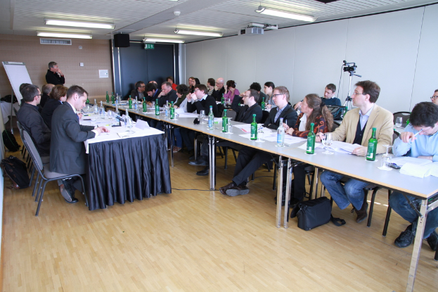 Ein Büroraum mit Mitarbeitern an Tischen, Rucksäcke auf dem Boden, ein Mann im Hintergrund und verschiedene Gegenstände wie Flaschen, Papiere, eine Kamera, Lautsprecher, Schilder, Projektor und Lampen.