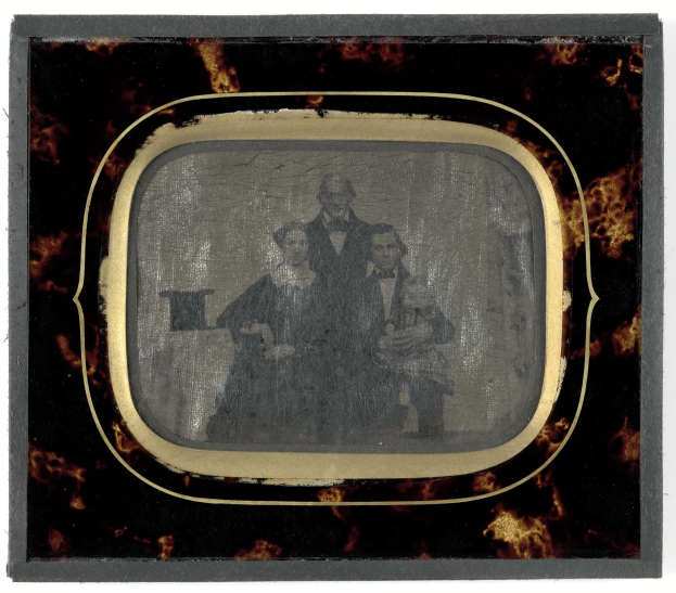 Altes Schwarz-Weiß-Foto eines Mannes in Anzug und Krawatte und einer Frau in unbekannter Kleidung nebeneinanderstehend mit ernsten Gesichtern vor einem Fernseher vor einem weißen Hintergrund.