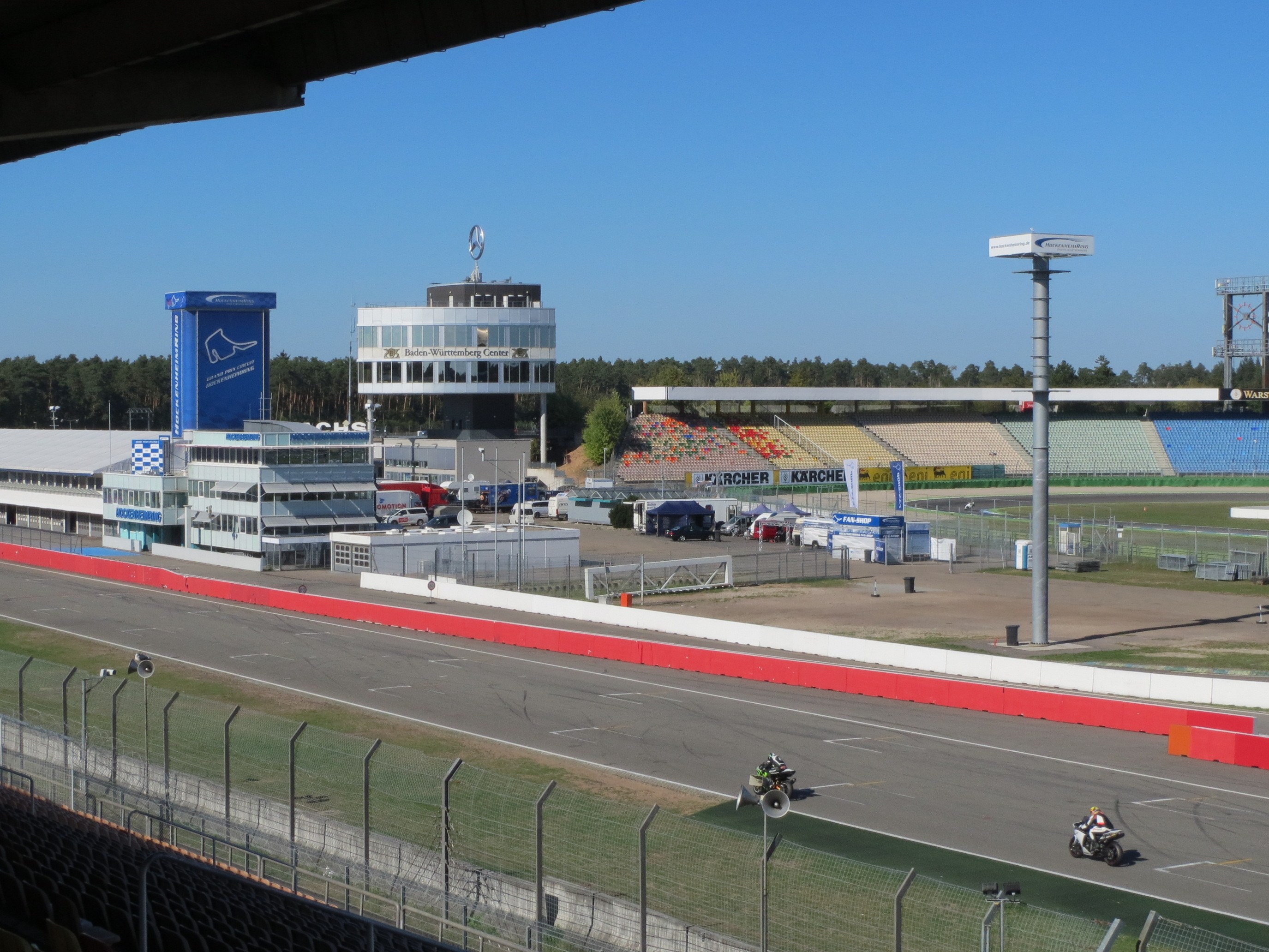 Gruppe von Motorradfahrern auf einer Rennstrecke, umgeben von einer Absperrung, Fahrzeugen, einem Gebäude, einem Schild, einer Tribüne und Bäumen unter einem bewölkten Himmel.