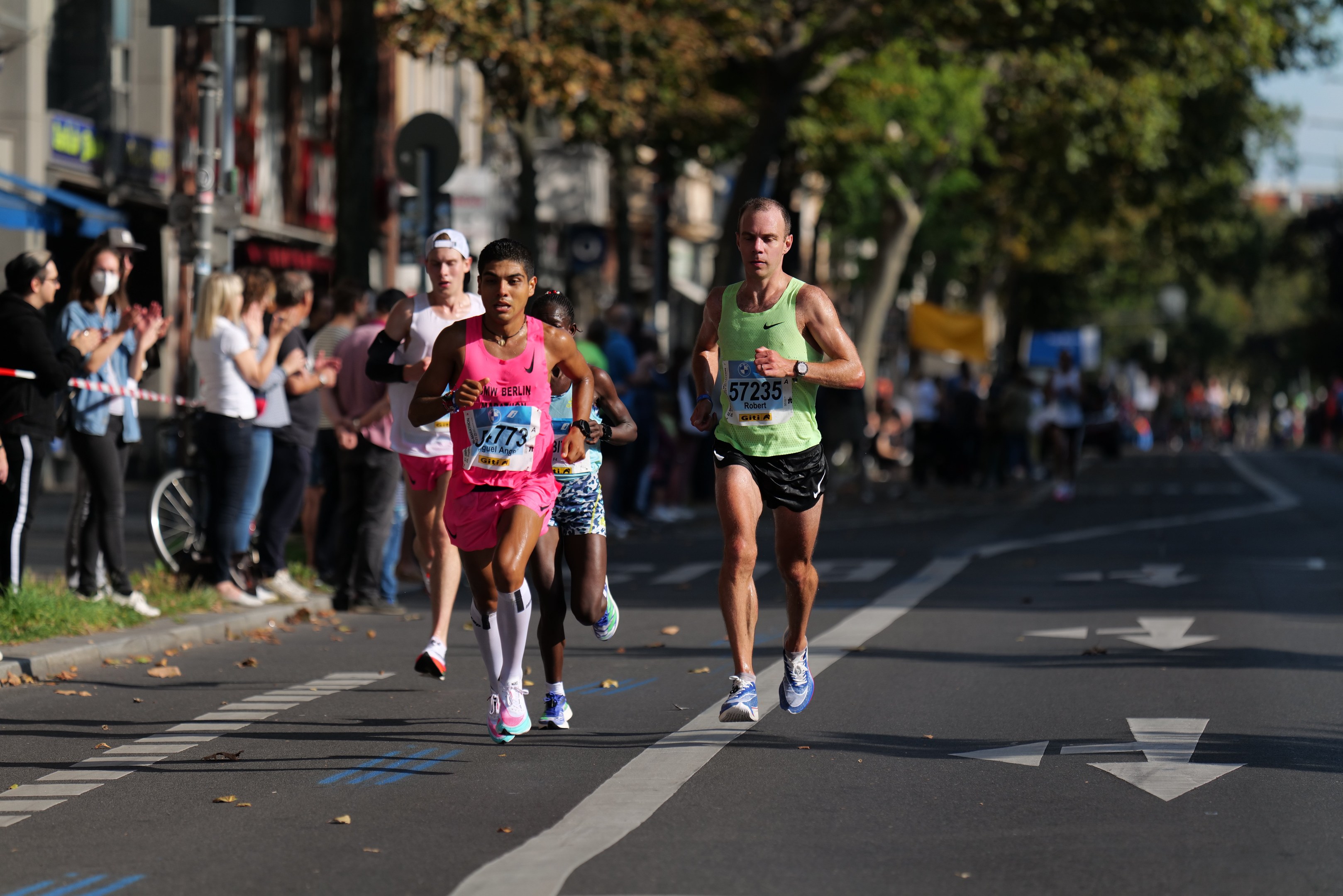 Gruppe von Menschen, die bei einem Marathon auf einer Stadtstraße laufen, mit Zuschauern auf der linken Seite, unscharfen Hintergrund mit Bäumen, Gebäuden, Pfählen, Schildern und einem Fahrrad.