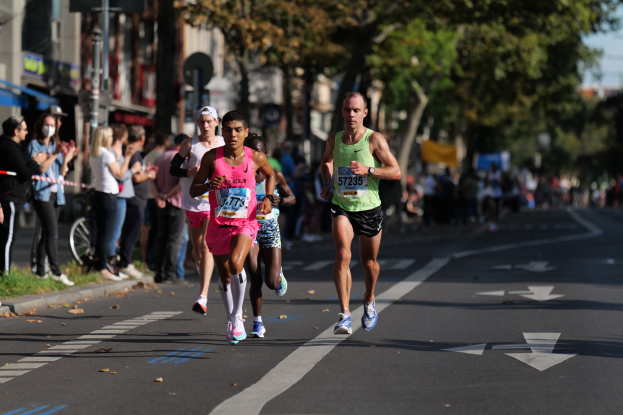 Gruppe von Menschen, die bei einem Marathon auf einer Stadtstraße laufen, mit Zuschauern auf der linken Seite, unscharfen Hintergrund mit Bäumen, Gebäuden, Pfählen, Schildern und einem Fahrrad.