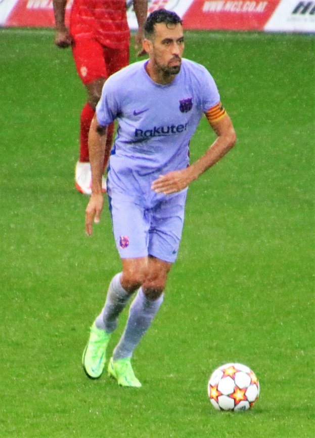 Fußballspieler in Sportbekleidung und Fußballschuhen auf einem Feld mit einem Textschild im Hintergrund.