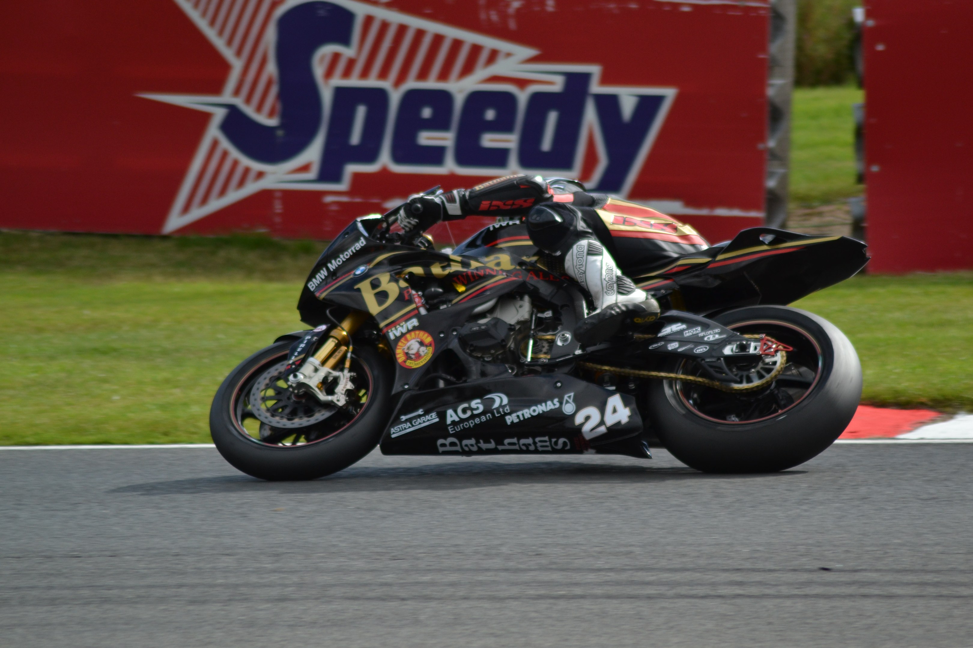 Eine Person in Helm und Schuhwerk fährt ein Motorrad auf einer Rennstrecke, die von Gras umgeben ist, mit einer Tafel mit Text im Hintergrund.