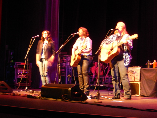 Drei Frauen treten auf einer Musikb├╝hne auf, jede singt in ein Mikrofon und spielt eine Gitarre, mit einem Tisch voller Wasserflaschen und zus├Ątzlichen Gitarren dahinter.