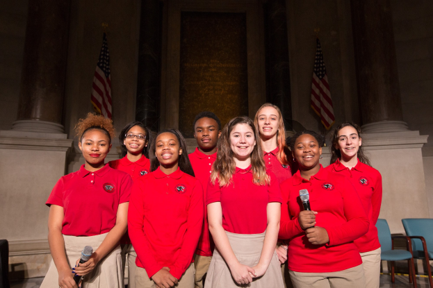 Gruppe junger Frauen in roten Shirts, die strahlend Mikrofone halten und neben Stühlen, Säulen und Fahnen in einem Saal stehen.