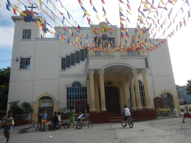 Eine Gruppe von Menschen, die Fahrräder vor einer weißen Kirche mit architektonischen Details wie Fenstern, Säulen, Bögen und einem Kreuzsymbol fahren, mit Pflanzen, einem Zaun, einer Fahne, Bäumen und einem bewölkten Himmel im Hintergrund und bunten Fahnen, die an einem Seil über der Kirche befestigt sind.