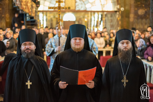 Drei Priester in schwarzen Gewändern und Kappen stehen vor einer Menge in einer Kirche, einer hält ein Buch, mit einem unscharfen Hintergrund und einem Wasserzeichen in der rechten unteren Ecke.