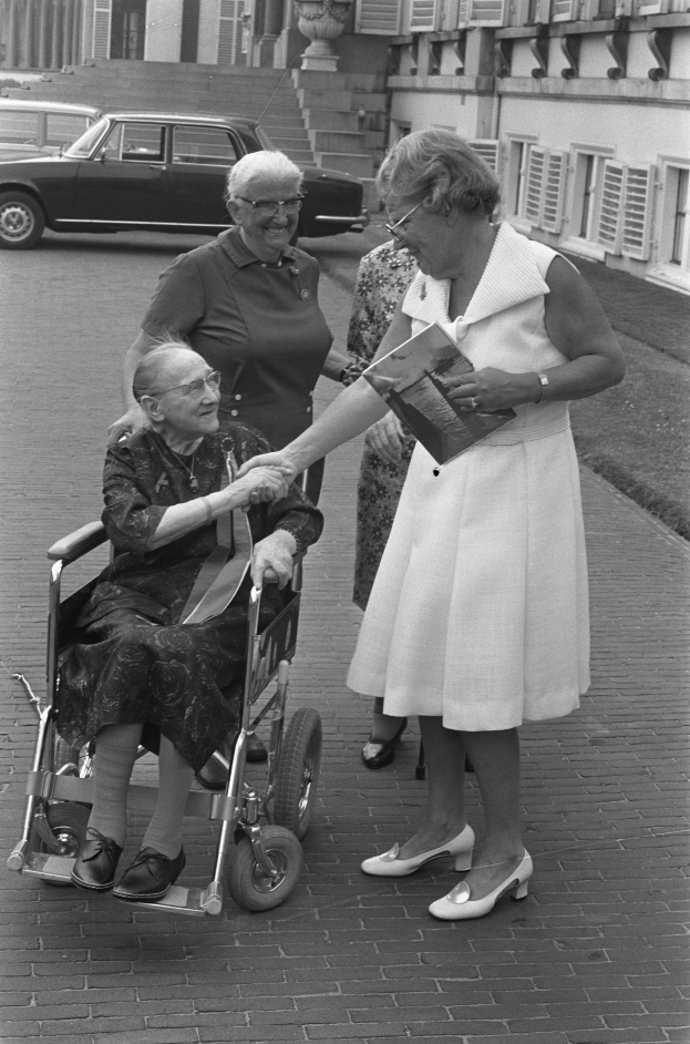 Eine Seniorin im Rollstuhl, die ein Kleid tr├Ągt und ein Buch h├Ąlt, wird von zwei Frauen mit Brille begr├╝├čt, mit einem Auto und einem Geb├Ąude im Hintergrund.
