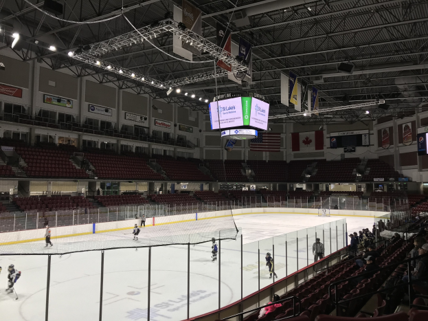 Eishockeyspiel in einer großen Arena mit einem zentralen Netz, Zuschauern auf Stühlen, Deckenbeleuchtung und Trägern sowie Bannern an den Wänden, die "St. Louis Blues Arena" beschriftet sind.