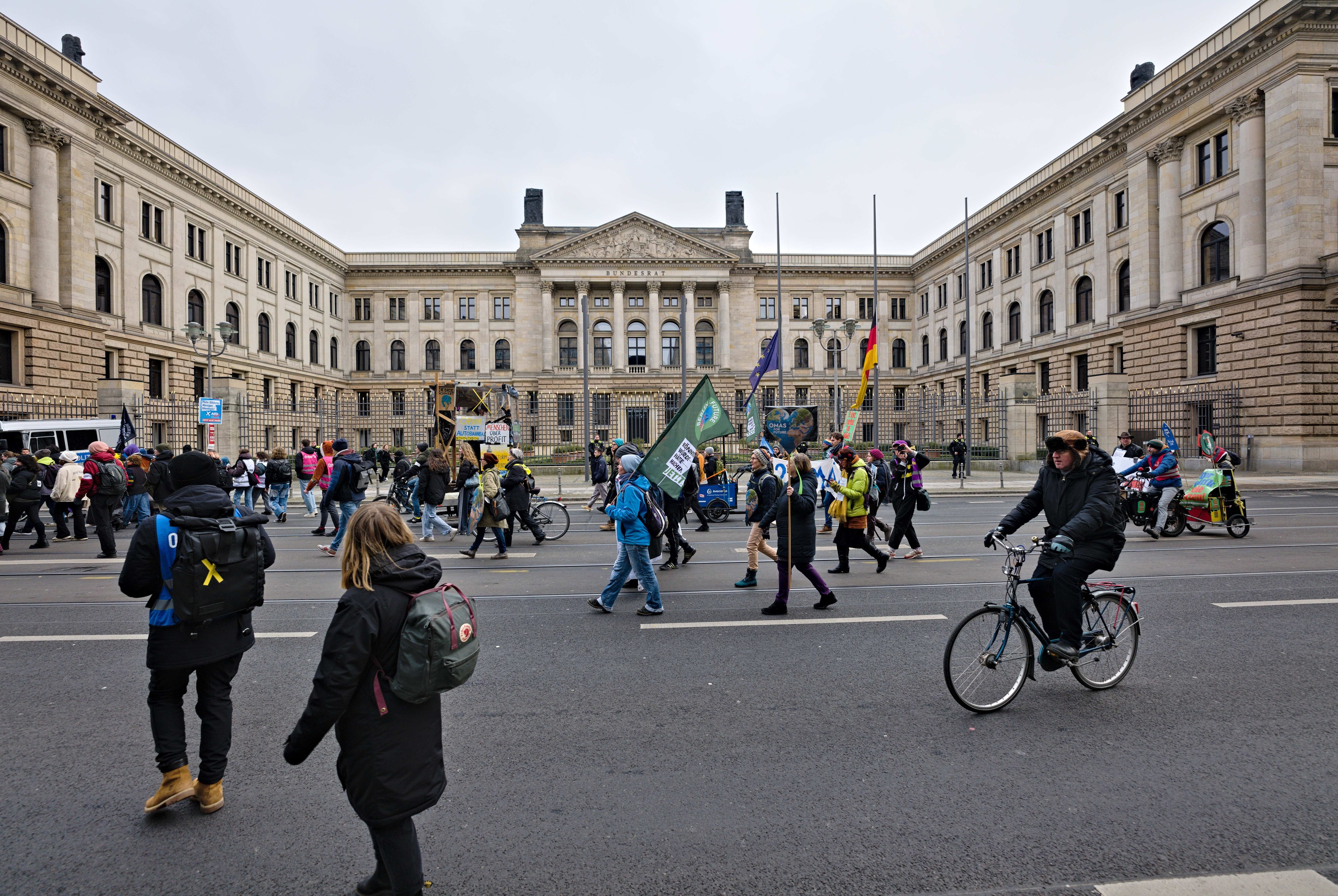 Eine Gruppe von Menschen geht vor einem großen Gebäude mit Fenstern, Säulen und Bögen die Straße entlang, einige tragen Taschen und Fahnen, andere fahren Fahrräder, im Hintergrund sind Fahrzeuge und Laternenmasten zu sehen.